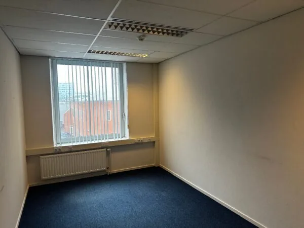 Lege kantoorruimte aan de Van Bleyswijckstraat met blauw tapijt, witte muren en een raam met verticale lamellen.